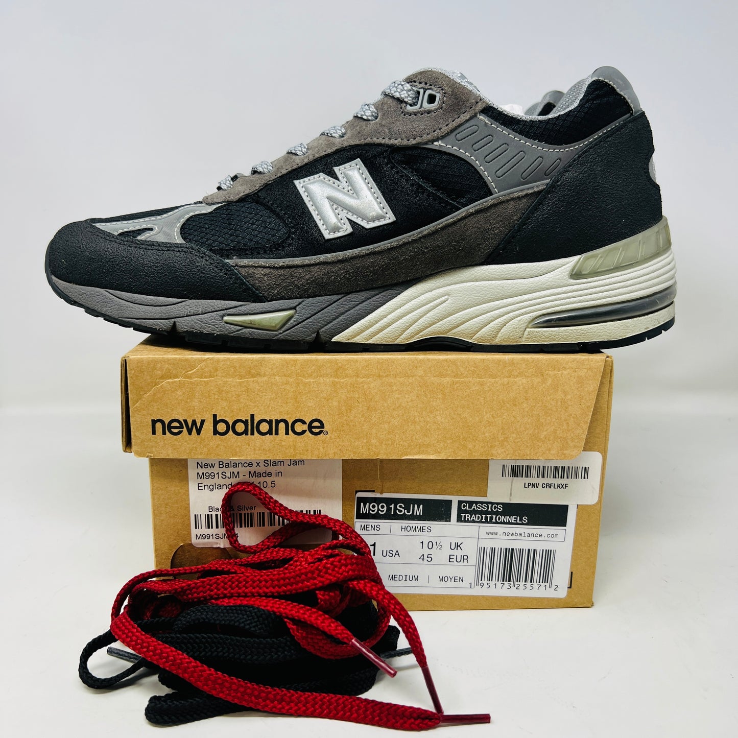 A New Balance 991 Slam Jam sneaker, in black and gray, sits on its box with tags attached. Red and black extra laces are neatly displayed in front, ready to refresh its look.