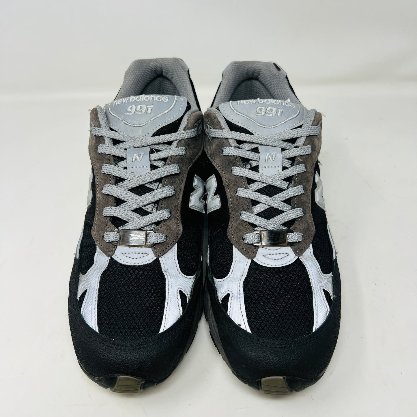 A New Balance 991 Slam Jam sneaker, in black and gray, sits on its box with tags attached. Red and black extra laces are neatly displayed in front, ready to refresh its look.