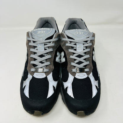 A New Balance 991 Slam Jam sneaker, in black and gray, sits on its box with tags attached. Red and black extra laces are neatly displayed in front, ready to refresh its look.