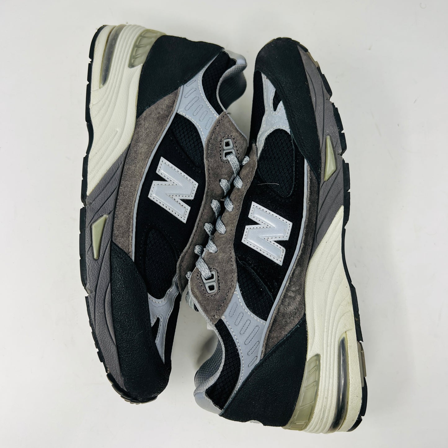 A New Balance 991 Slam Jam sneaker, in black and gray, sits on its box with tags attached. Red and black extra laces are neatly displayed in front, ready to refresh its look.