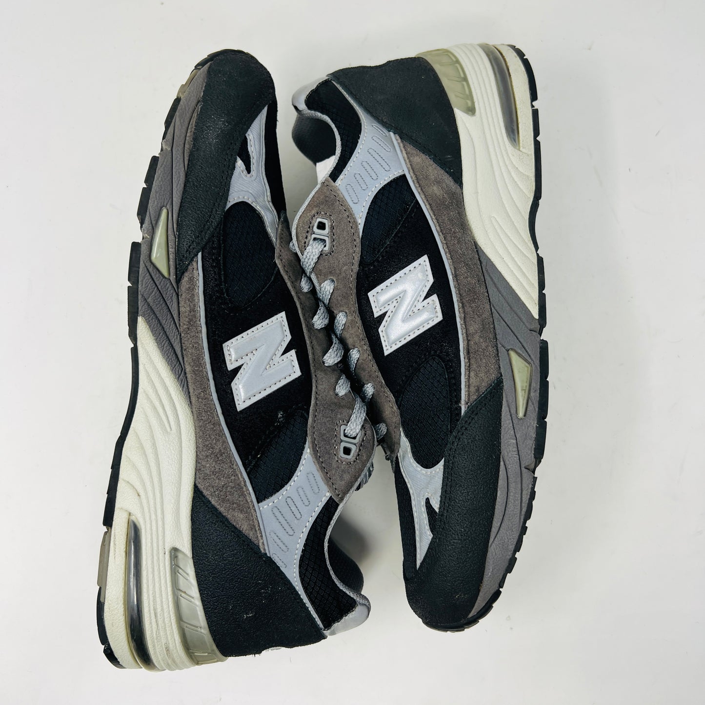 A New Balance 991 Slam Jam sneaker, in black and gray, sits on its box with tags attached. Red and black extra laces are neatly displayed in front, ready to refresh its look.