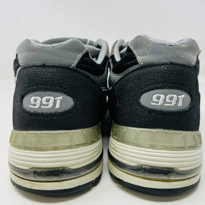 A New Balance 991 Slam Jam sneaker, in black and gray, sits on its box with tags attached. Red and black extra laces are neatly displayed in front, ready to refresh its look.