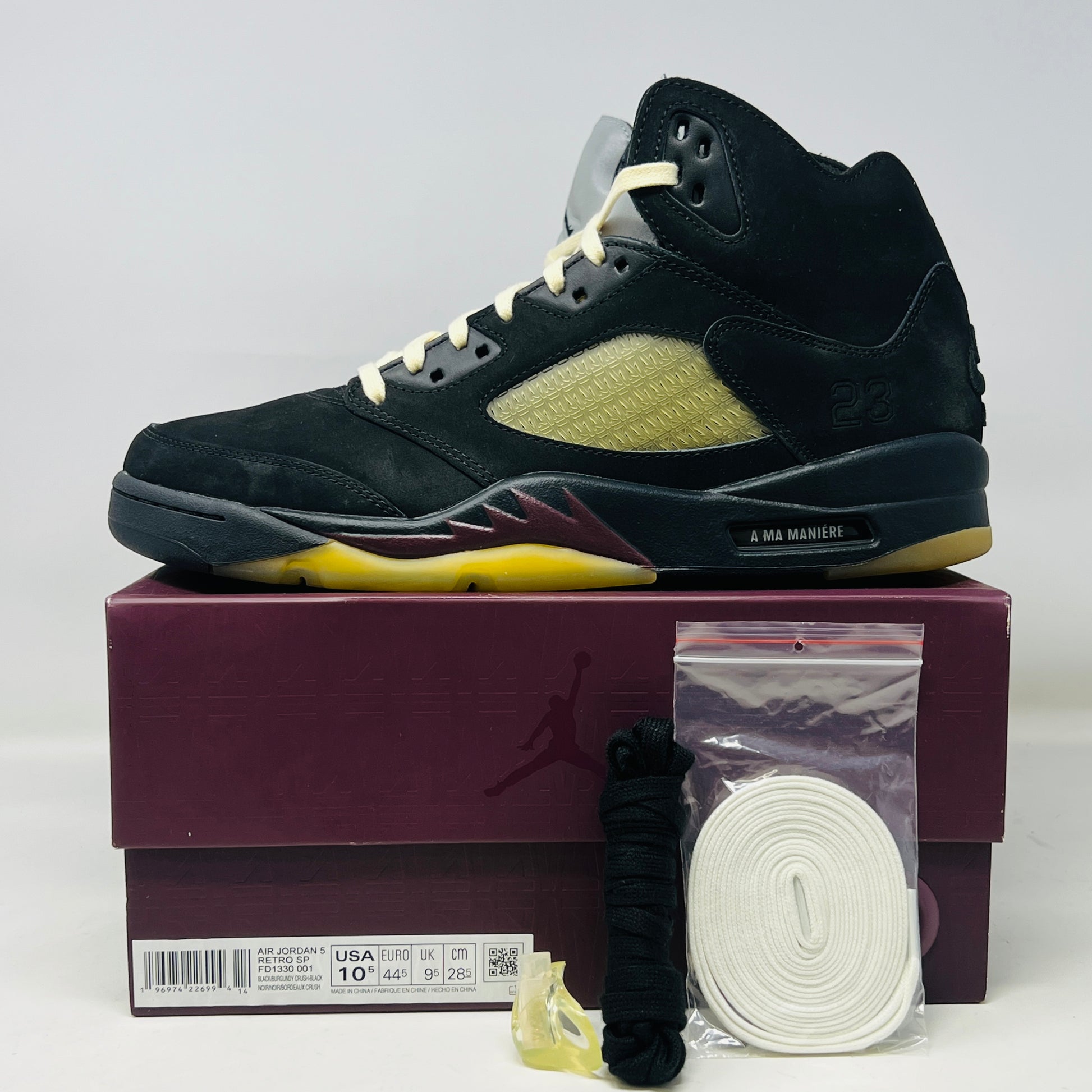 The Jordan 5 Retro A Ma Maniére Dusk sneaker, featuring sleek black and gold uppers and a translucent yellow sole, sits on a maroon shoebox. It includes black and white laces, extra pairs, and a yellow lace lock, all presented in front of the 2023 limited edition box from Jordan.
