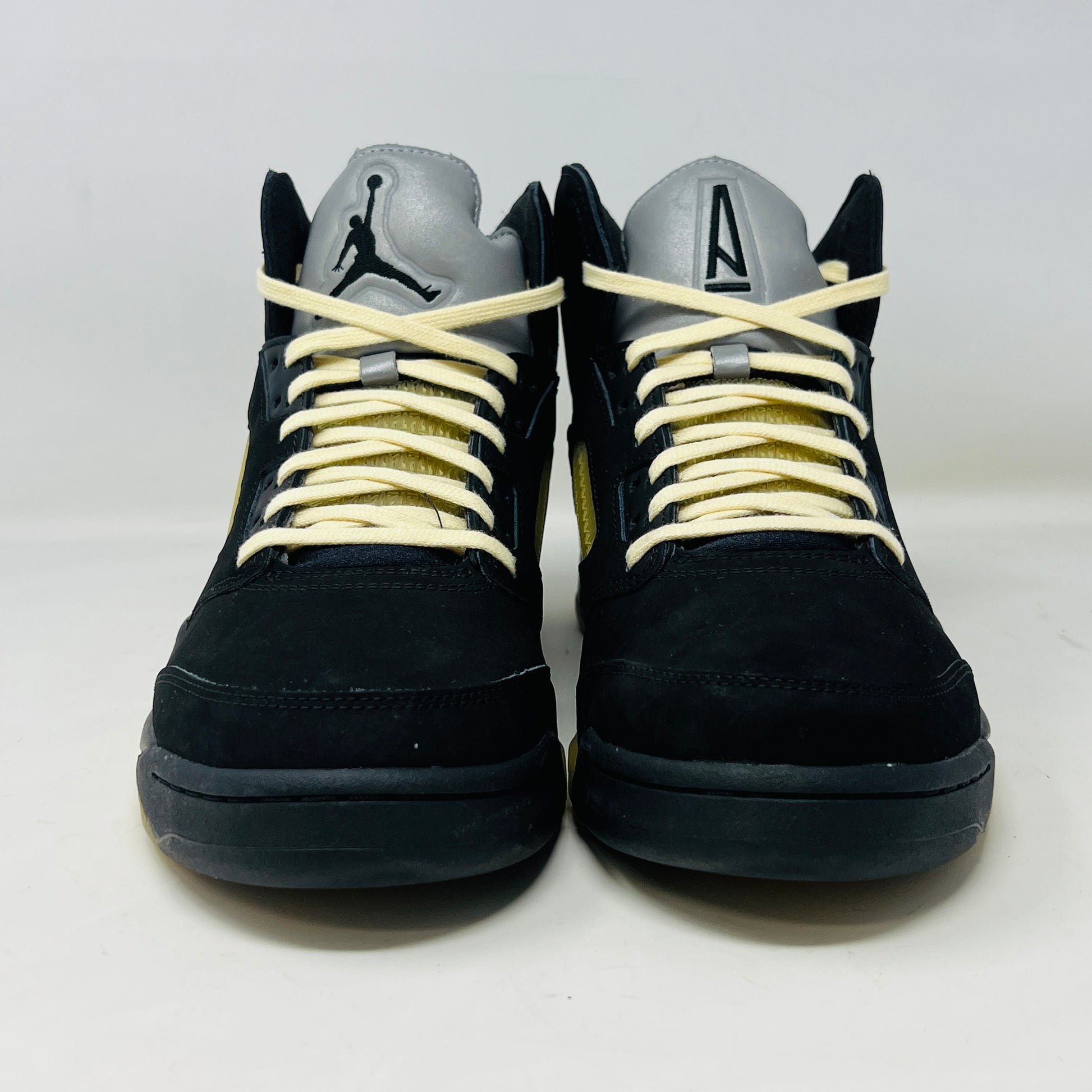 The Jordan 5 Retro A Ma Maniére Dusk sneaker, featuring sleek black and gold uppers and a translucent yellow sole, sits on a maroon shoebox. It includes black and white laces, extra pairs, and a yellow lace lock, all presented in front of the 2023 limited edition box from Jordan.