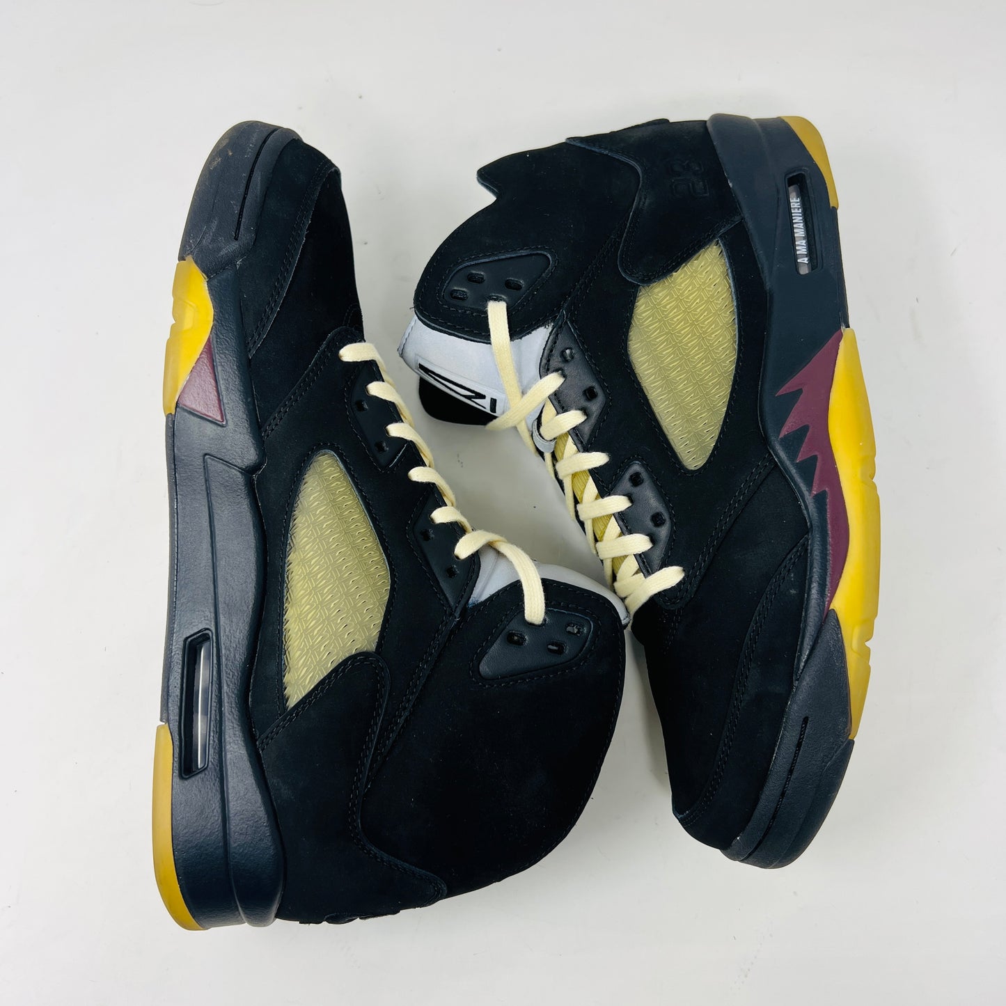 The Jordan 5 Retro A Ma Maniére Dusk sneaker, featuring sleek black and gold uppers and a translucent yellow sole, sits on a maroon shoebox. It includes black and white laces, extra pairs, and a yellow lace lock, all presented in front of the 2023 limited edition box from Jordan.