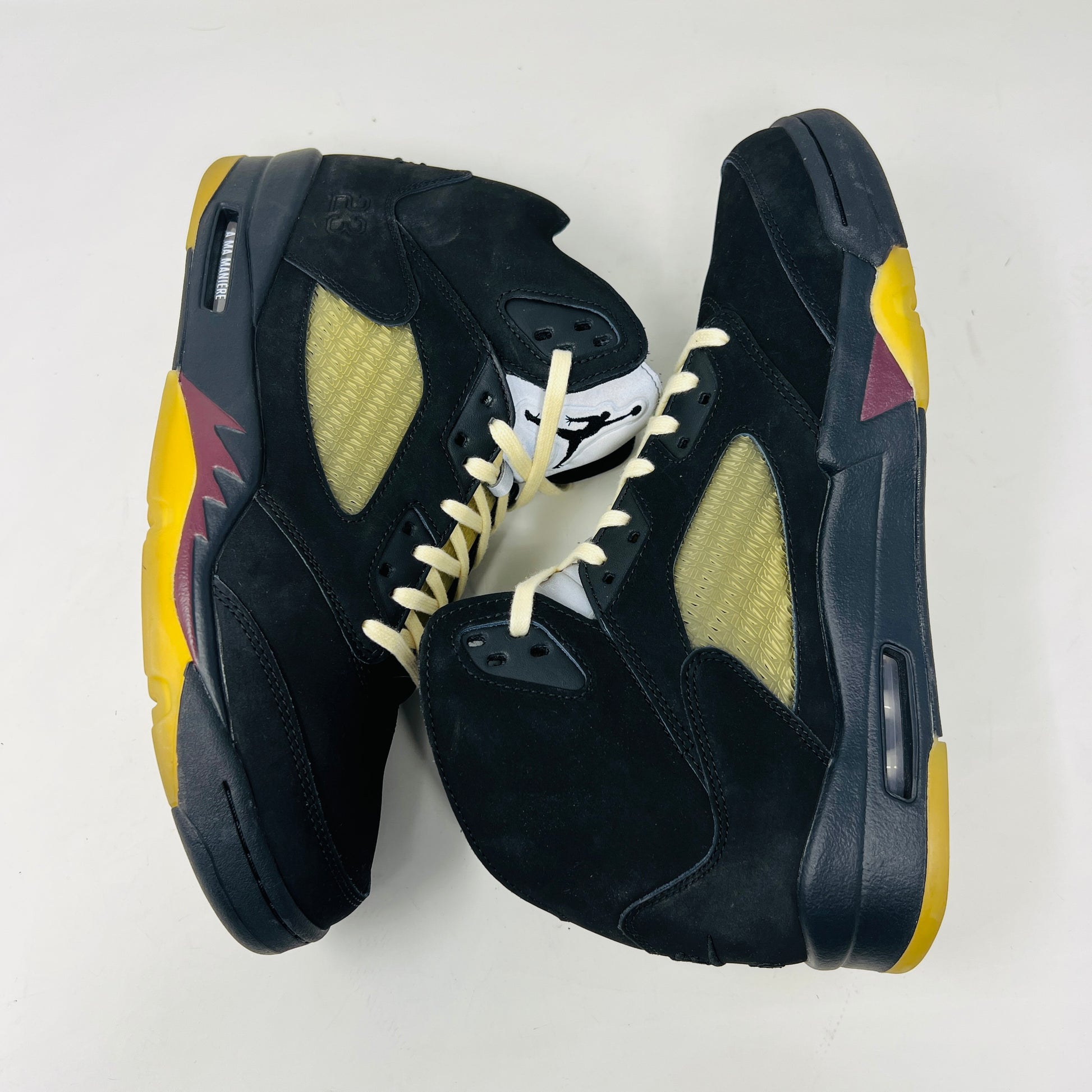 The Jordan 5 Retro A Ma Maniére Dusk sneaker, featuring sleek black and gold uppers and a translucent yellow sole, sits on a maroon shoebox. It includes black and white laces, extra pairs, and a yellow lace lock, all presented in front of the 2023 limited edition box from Jordan.