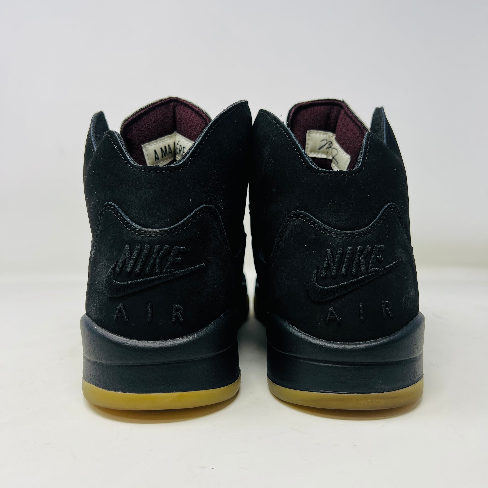 The Jordan 5 Retro A Ma Maniére Dusk sneaker, featuring sleek black and gold uppers and a translucent yellow sole, sits on a maroon shoebox. It includes black and white laces, extra pairs, and a yellow lace lock, all presented in front of the 2023 limited edition box from Jordan.