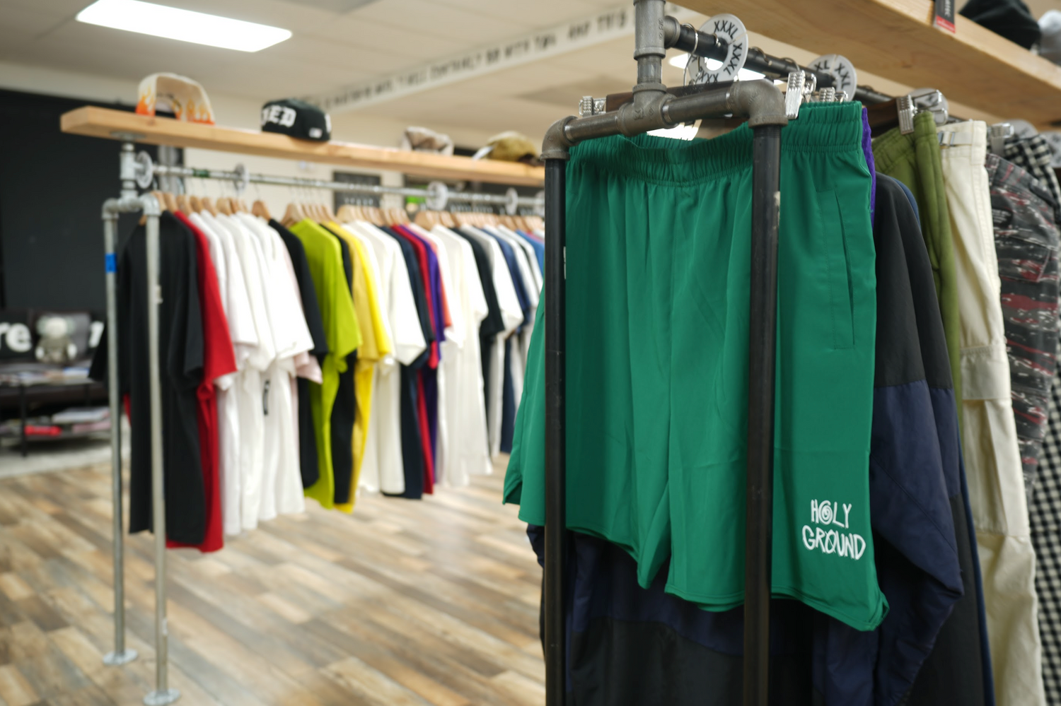 A clothing store interior featuring a rack of colorful t-shirts on wooden hangers. In the foreground, green shorts with HOLY GROUND written on them hang from a metal pipe rack. Wooden flooring and bright lighting complete the scene.