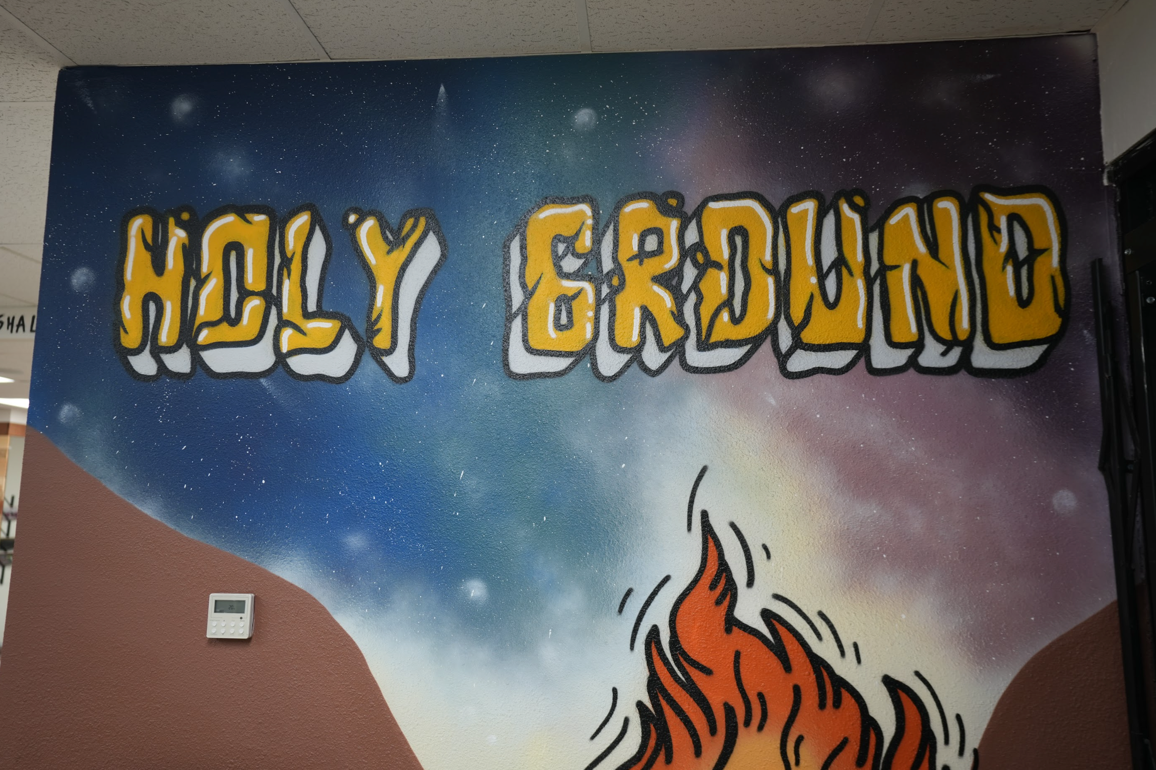 A mural on a wall with a cosmic background and the words HOLY GROUND in bold, yellow letters. Below, there is a stylized illustration of a fire with red and orange flames. A thermostat is visible on the left side of the wall.