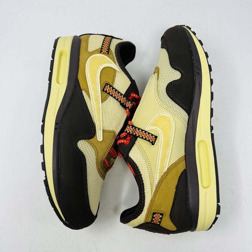 A brand new Nike Air Max 1 TS Cactus Jack Baroque Brown sneaker in yellow and brown with orange accents sits on a brown Nike shoebox. It boasts a wavy design, textured fabric, extra brown laces, and a visible air unit in the sole against a plain white background.