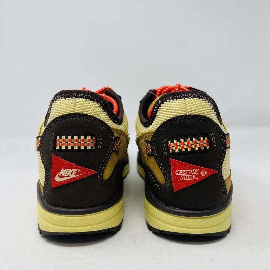 A brand new Nike Air Max 1 TS Cactus Jack Baroque Brown sneaker in yellow and brown with orange accents sits on a brown Nike shoebox. It boasts a wavy design, textured fabric, extra brown laces, and a visible air unit in the sole against a plain white background.