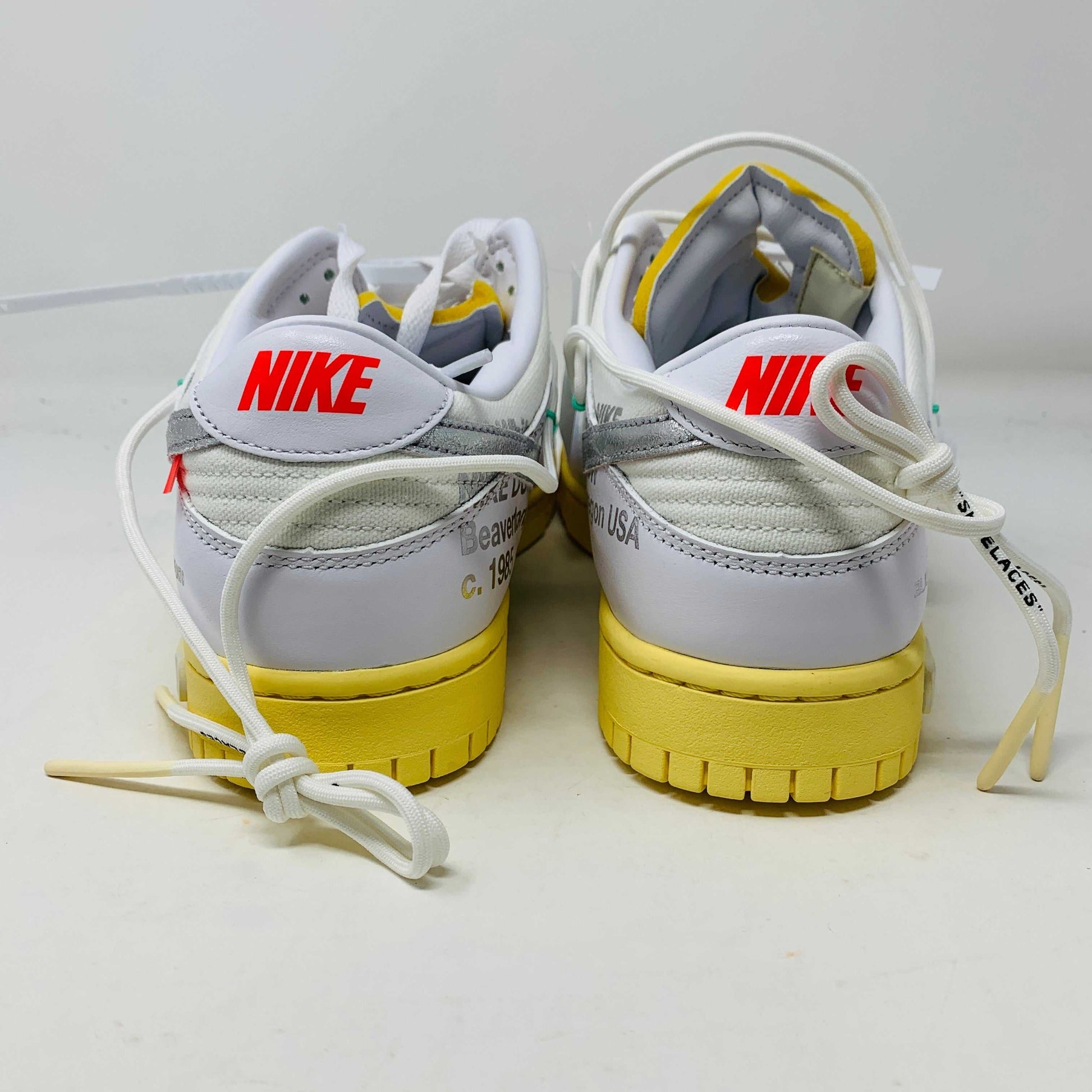 The Nike Dunk Low Off White Lot 1 sneaker, white with a metallic silver swoosh and vibrant accents, rests on a brown shoebox. Its unique lace design and 01 of 50 detailing highlight its 2021 release against a plain white backdrop.