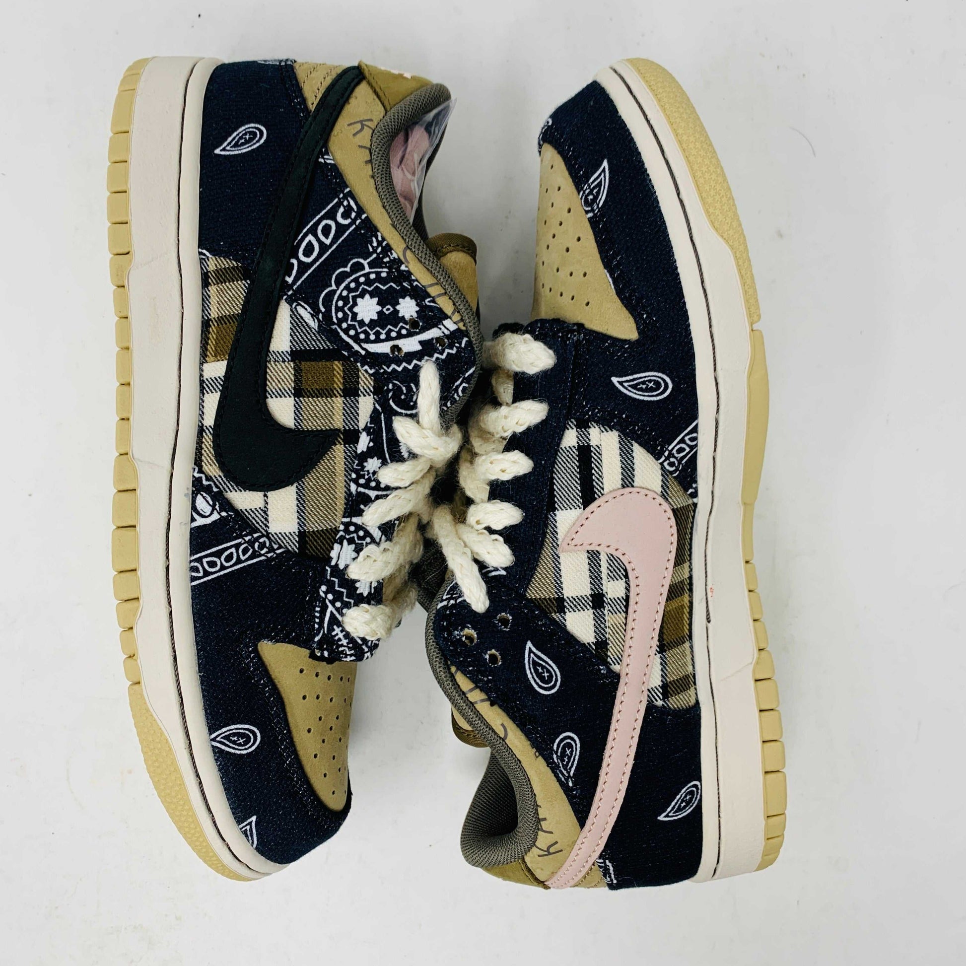 The Nike SB Dunk Low Travis Scott sneakers feature beige suede toe caps, black bandana-patterned overlays, white rope laces, authentic pink accents, and a brown label with text on the tongue. Brand new with extra laces, the interior is visible at the top.