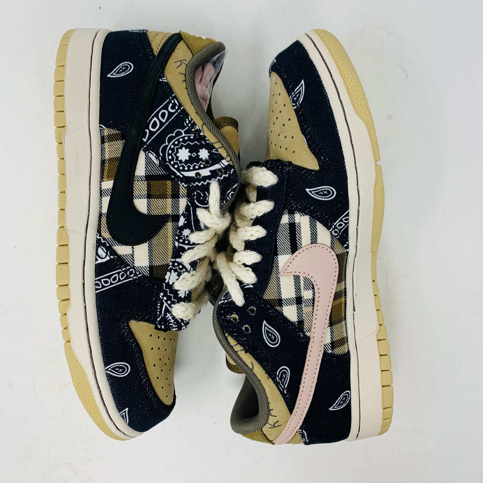 The Nike SB Dunk Low Travis Scott sneakers feature beige suede toe caps, black bandana-patterned overlays, white rope laces, authentic pink accents, and a brown label with text on the tongue. Brand new with extra laces, the interior is visible at the top.