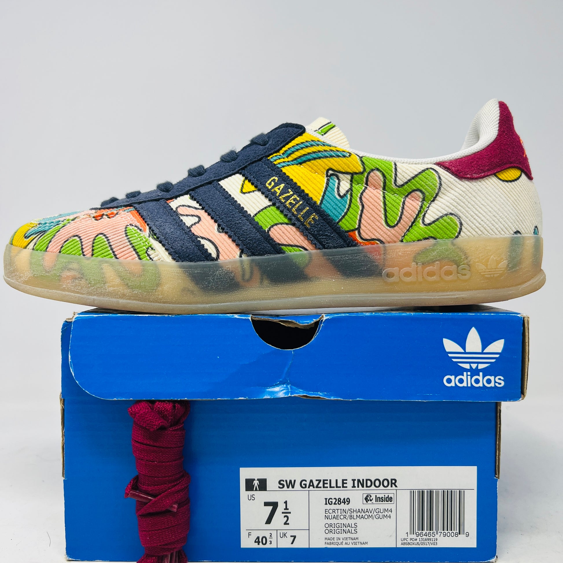 The Adidas Gazelle Indoor Sean Wotherspoon sneaker, with colorful abstract uppers in yellow, red, pink, and green, dark blue stripes, red laces, and translucent soles, rests on a blue box showing size and product details.