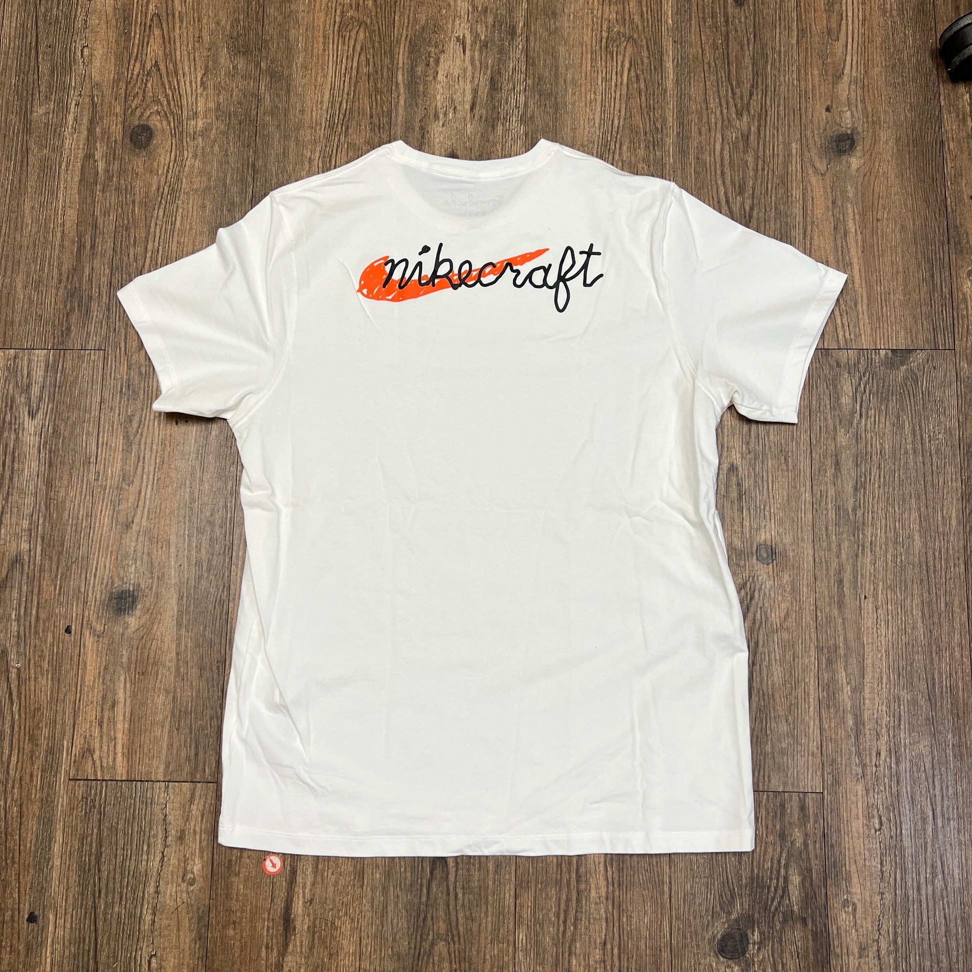 A brand new Nike Tom Sachs - Nikecraft studio shirt in white lies flat on a wooden floor, featuring a small logo with text on the left chest and another logo inside the back neckline.