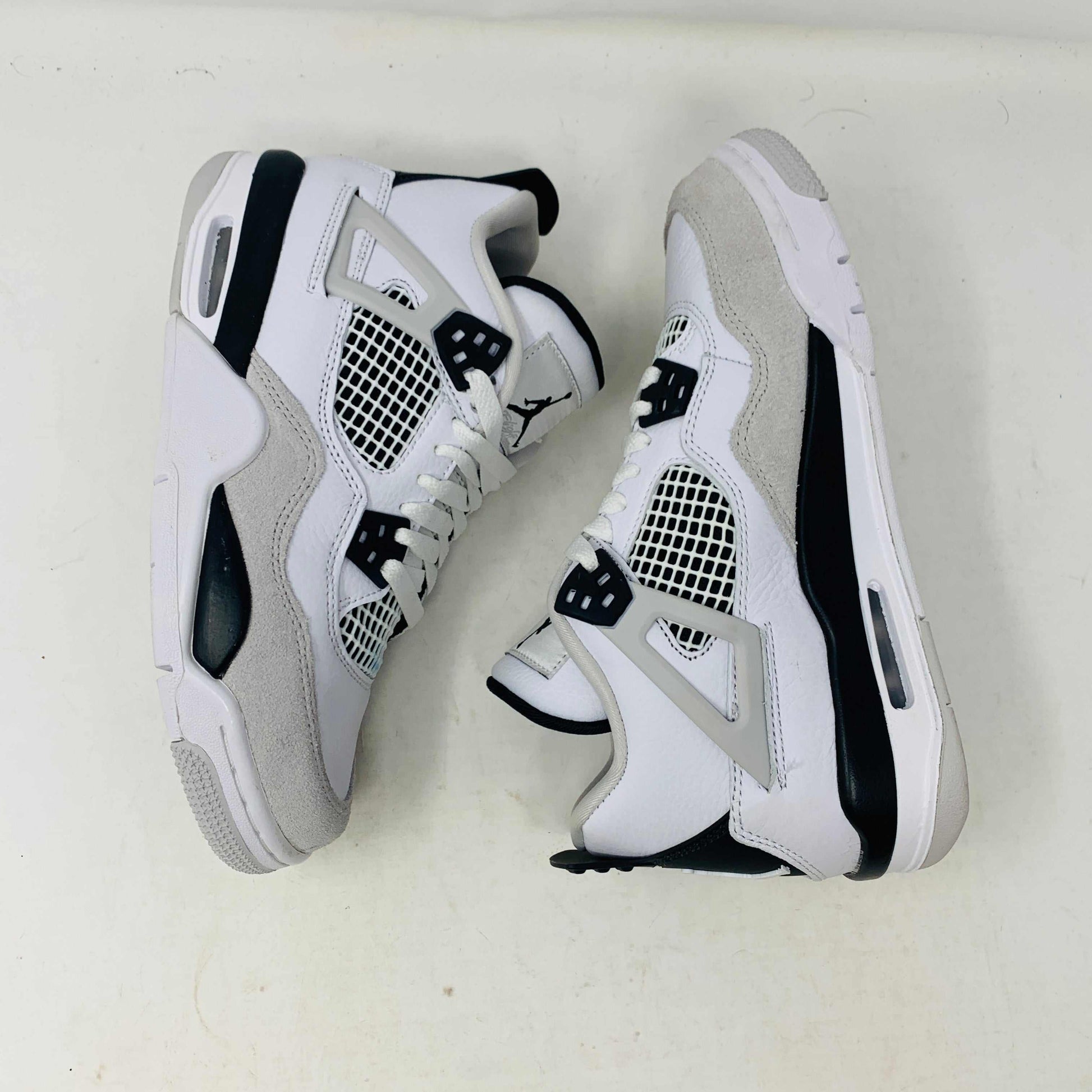 The Jordan 4 Military Black GS sneakers, featuring distinctive side netting and a logo on the tongue, are brand new and authentic in white and gray with black accents. They face forward on a white surface against a plain background, embodying modern style.
