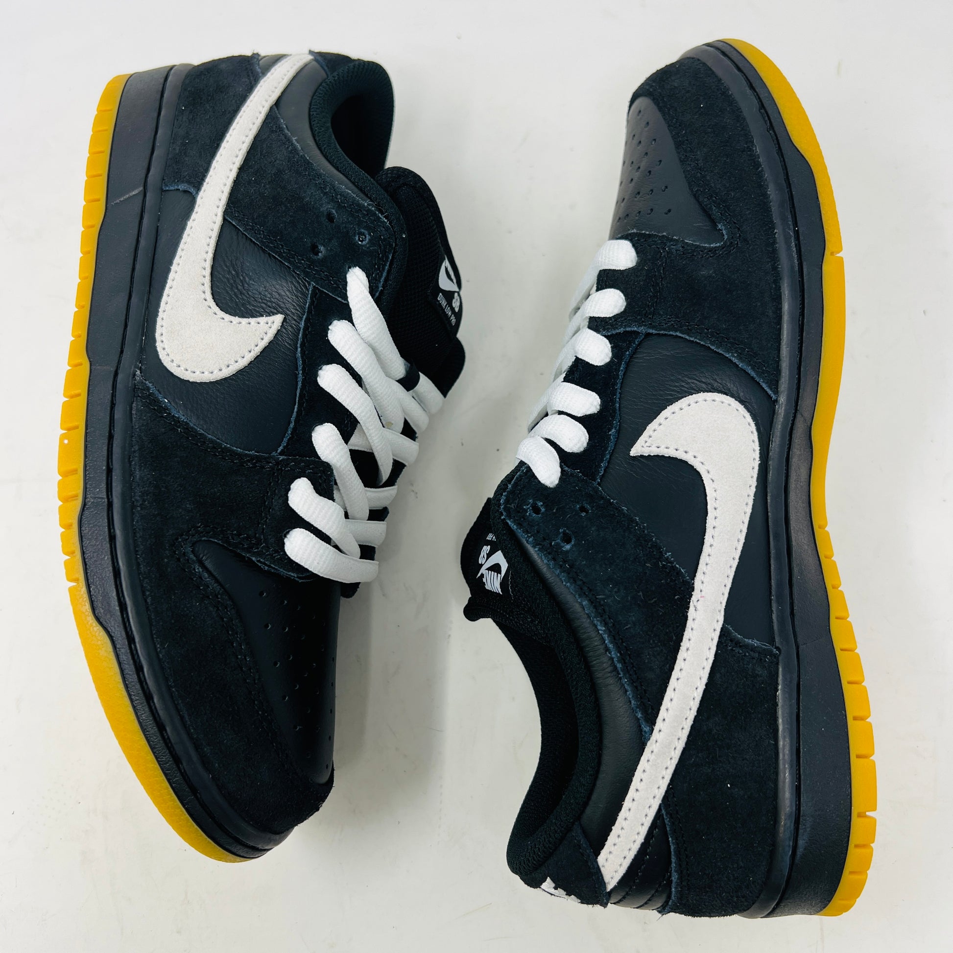 A black Nike Dunk Low Pro SB Black White Gum sneaker with white laces and swoosh sits on a purple Nike SB box. The 2025 shoe has a yellow sole, leather-texture finish, and both are in new condition.
