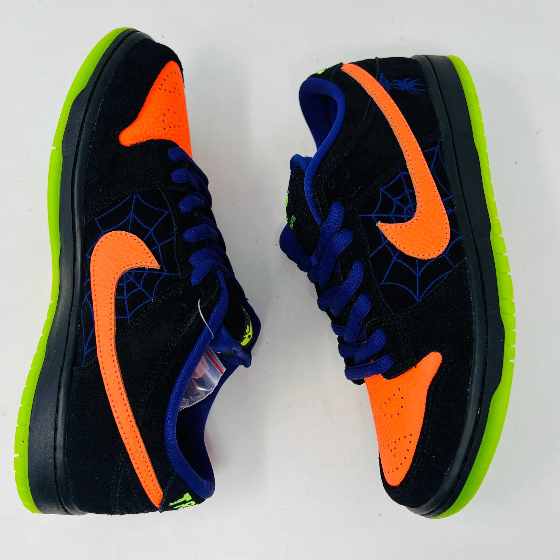 The Nike SB Dunk Low Night of Mischief sneakers feature Halloween-themed designs with orange swooshes, green soles, and spider web details. They come in a coffin-shaped box adorned with spooky jack-o-lanterns and webs, resting on a wooden floor.