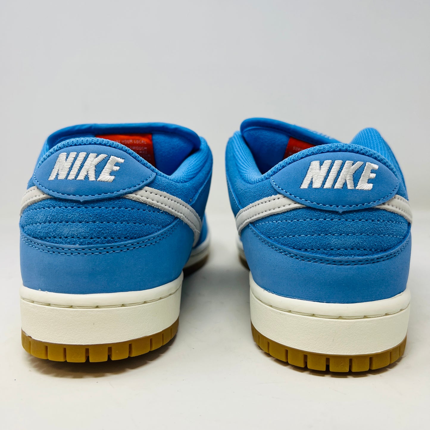 The Nike SB Dunk Low Pro University Blue Gum sneaker, featuring a white swoosh, laces, midsole, and gum-colored outsole, is placed on a brown shoebox with an orange swoosh against a plain white background.