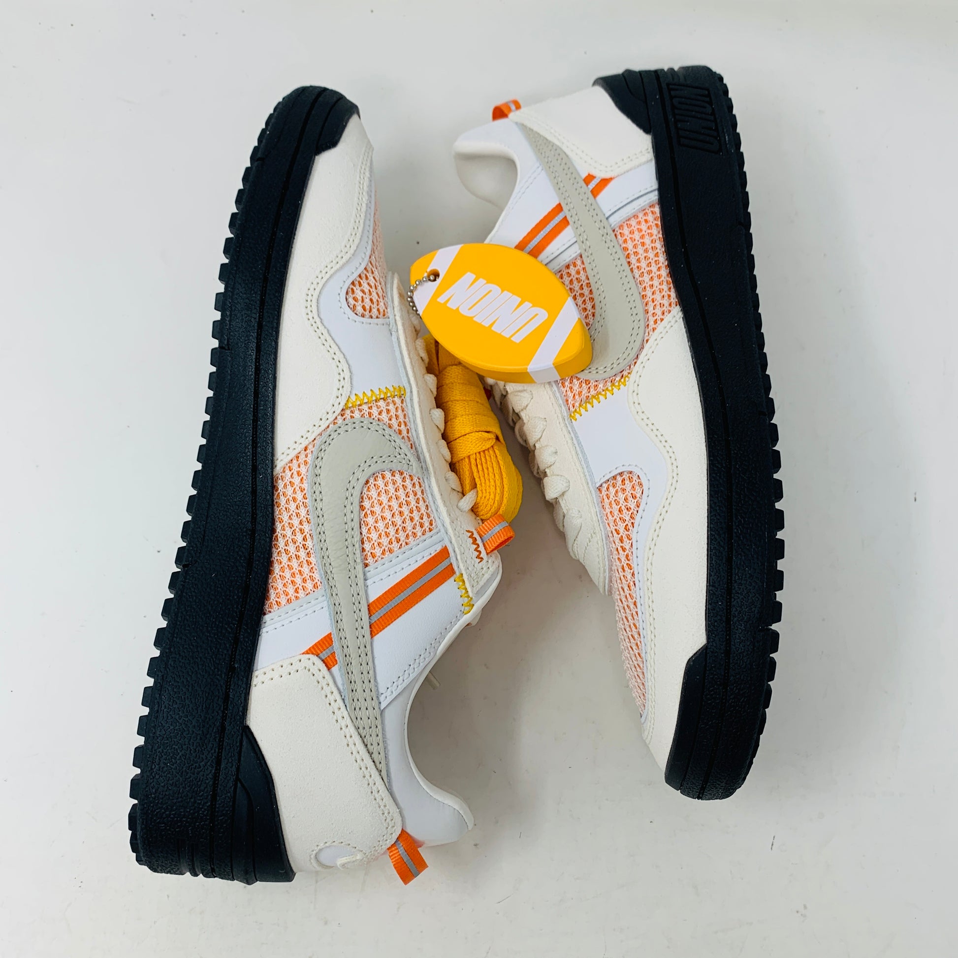 The pristine Nike Field General Union LA Ivory sneakers, in white and orange with black soles, rest on an orange box. Featuring a UNION tag, Nike logo, and a blue sticker with a silhouetted man, theyre perfect for any accessories collection.