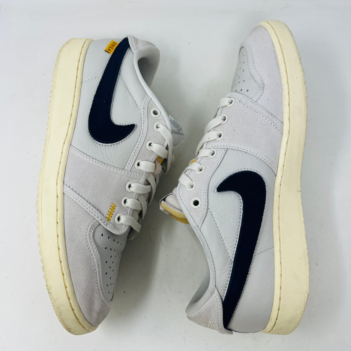 The Jordan 1 Retro AJKO Low SP Union Sail Leather sneakers feature black swooshes, subtle yellow stitching, and white laces. Pictured on a pristine white background, they include extra laces for versatile styling.
