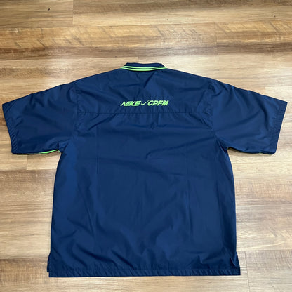 This brand new size Medium Nike x Cactus Plant Flea Market S/S Polo in navy and neon green features NIKE and CPEM on the front/sleeves, a collar zipper, and a small Nike logo, laid flat on wooden floor.