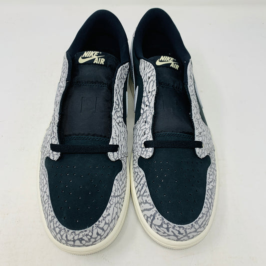 The Jordan 1 Retro Low OG Black Cement sneaker, with black and gray elephant print and a white swoosh, is showcased on a matching shoe box. A small brown box with extra laces is beside it, all set against a plain background.