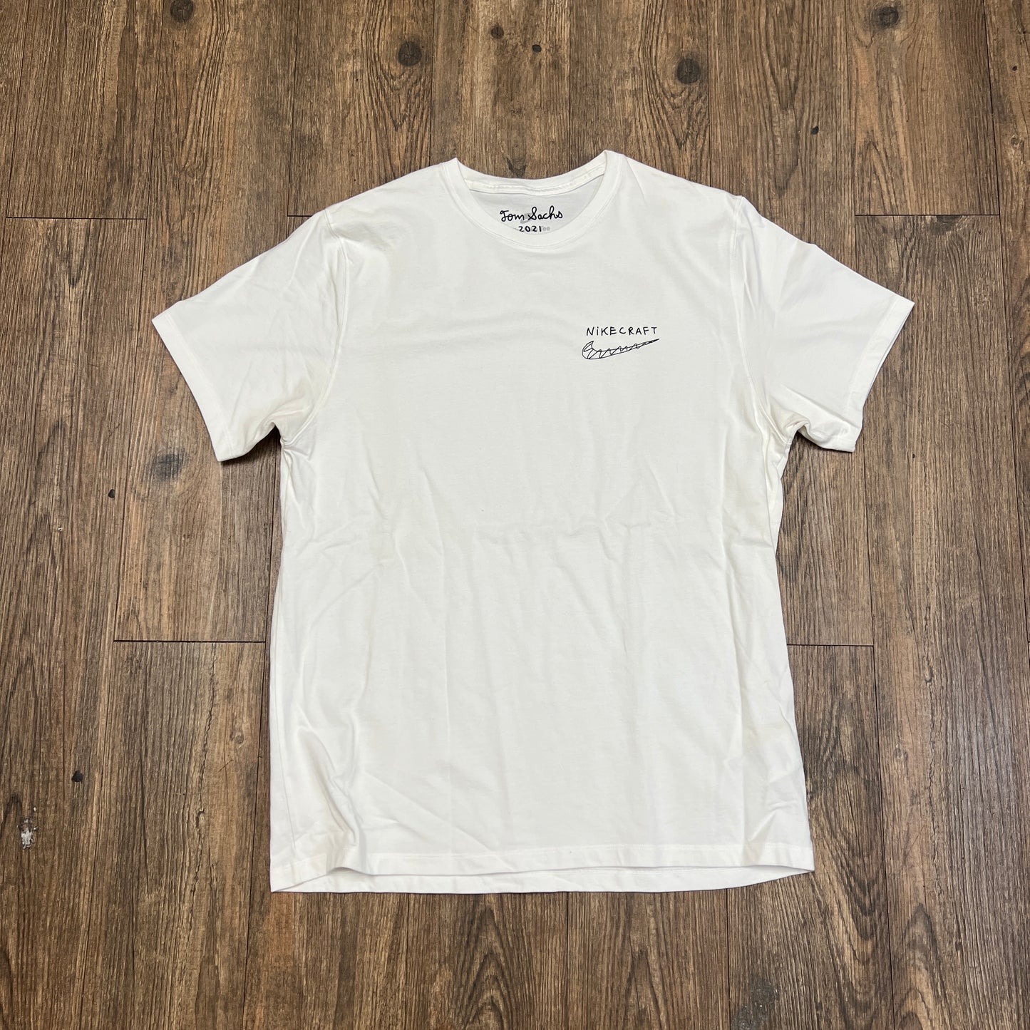 A brand new Nike Tom Sachs - Nikecraft studio shirt in white lies flat on a wooden floor, featuring a small logo with text on the left chest and another logo inside the back neckline.