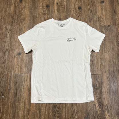 A brand new Nike Tom Sachs - Nikecraft studio shirt in white lies flat on a wooden floor, featuring a small logo with text on the left chest and another logo inside the back neckline.