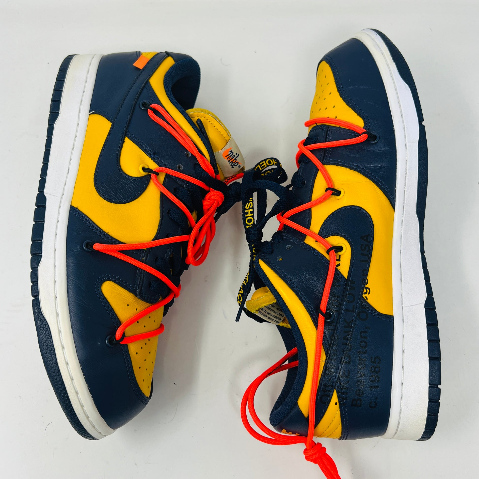 The 2019 Nike Dunk Low Off-White University Gold sneakers feature a navy and yellow design, accented with bold red laces and attached white tags and a zip tie. Displayed atop their box, the label lists them as US size 10.5 and UK size 12, all set against a plain white background.