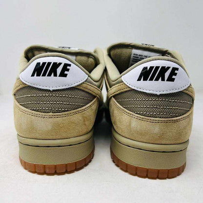 The Nike SB Dunk Low Pro B Parachute Beige, with a beige swoosh and gum sole, is displayed on a purple Nike SB box. New tan suede sneaker includes extra laces neatly rolled up in front against a plain white background.