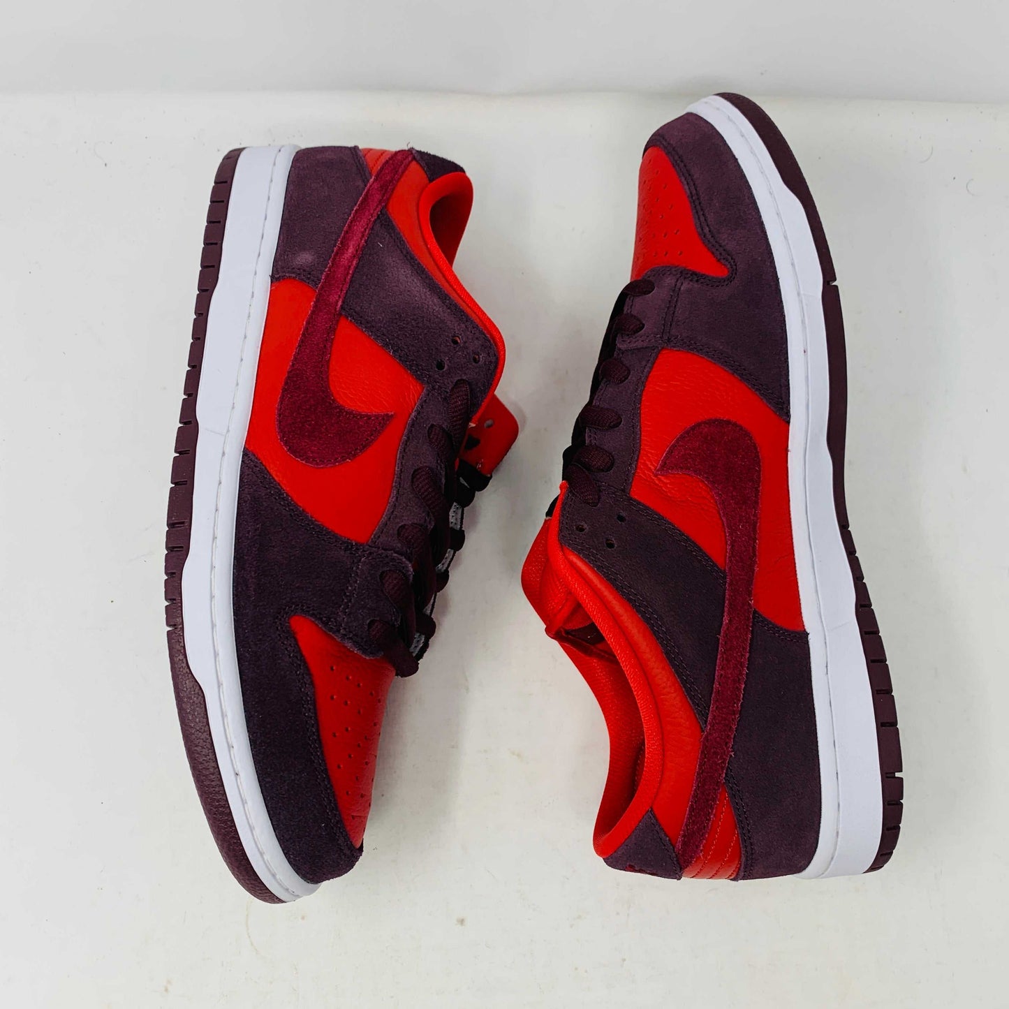A 100% authentic, brand new Nike SB Dunk Low Cherry sneaker in red and dark brown sits on a branded purple Nike SB shoebox. It features a red swoosh, toe box, dark brown overlays, extra laces, white midsole, and dark brown outsole.