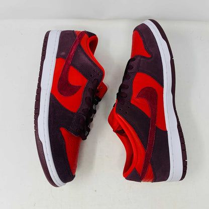 A 100% authentic, brand new Nike SB Dunk Low Cherry sneaker in red and dark brown sits on a branded purple Nike SB shoebox. It features a red swoosh, toe box, dark brown overlays, extra laces, white midsole, and dark brown outsole.