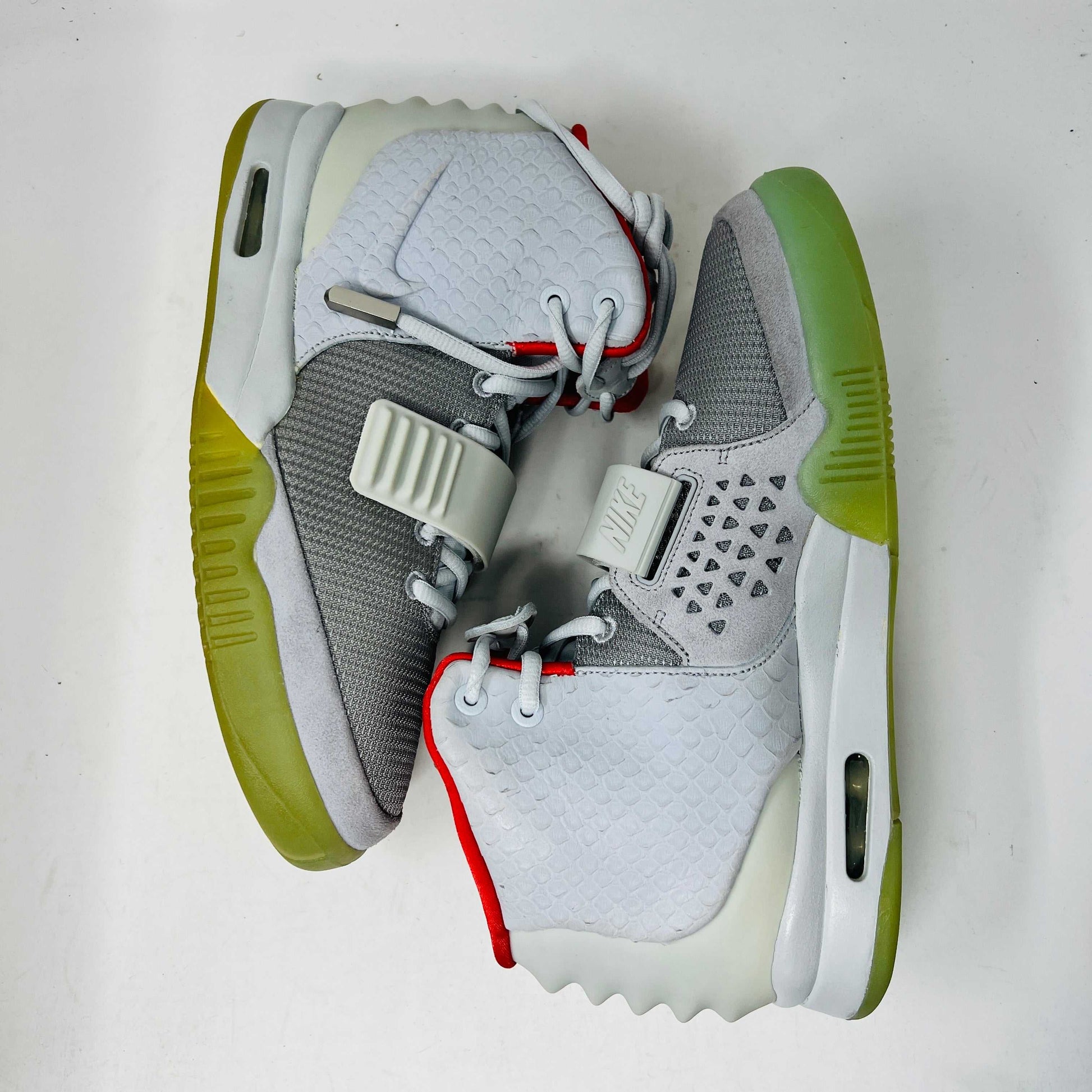 A pair of new Nike Air Yeezy 2 Pure Platinum high-tops rests on a cardboard box. The sneakers are mostly gray with a red lining, midfoot strap, and green-tinted soles. The label reads Air Yeezy 2 NRG, size 9, showcasing authentic detailing by Nike.