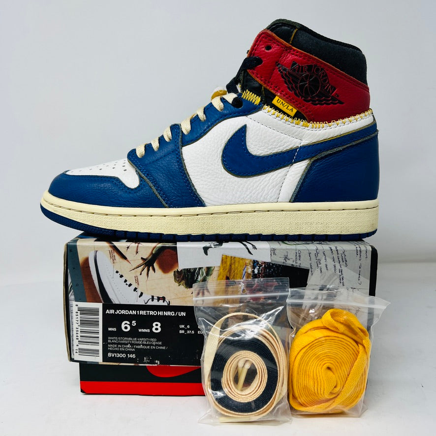 A close-up of a single Jordan 1 Retro High Union Los Angeles Storm Blue sneaker in white, blue, red, and black with clean uppers and yellow stitching. Its displayed on a Star Lost box with extra yellow and black laces packaged in front.