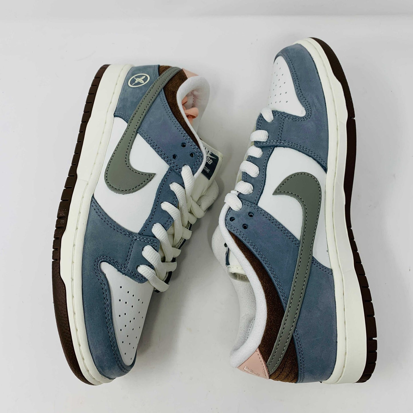 The Nike SB Dunk Low Yuto Horigome sneaker, with a white and light blue design featuring a gray swoosh, is displayed on a white Nike SB box. The authentic shoe has pink and brown laces, a brown sole, and an embroidered logo on the heel.