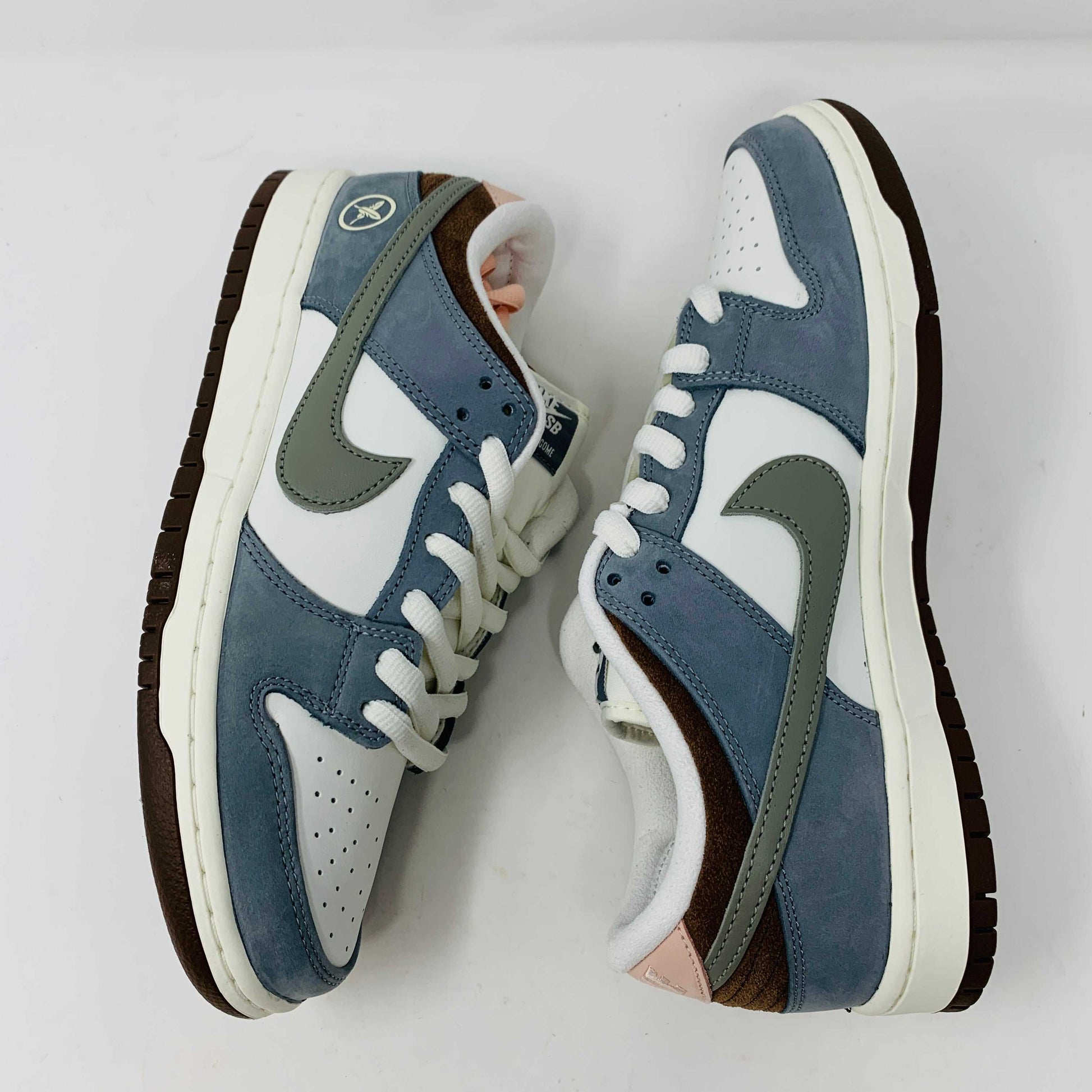 The Nike SB Dunk Low Yuto Horigome sneaker, with a white and light blue design featuring a gray swoosh, is displayed on a white Nike SB box. The authentic shoe has pink and brown laces, a brown sole, and an embroidered logo on the heel.