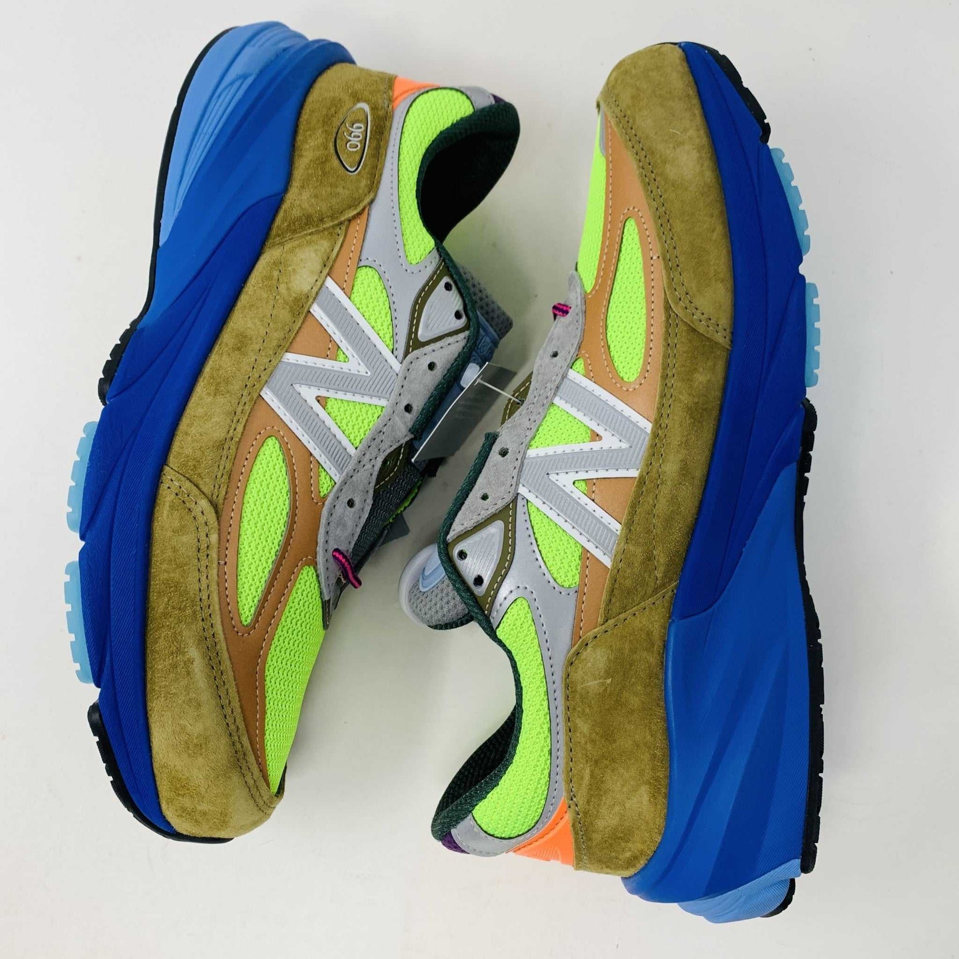 The New Balance 990v6 MiUSA Action Bronson Baklava sneaker, displayed on a white shoe box, boasts vibrant neon green, blue, and brown colors with suede and mesh materials. It features a distinctive design with a bold sole and branding on the side—truly authentic for 2023.