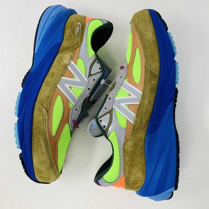 The New Balance 990v6 MiUSA Action Bronson Baklava sneaker, displayed on a white shoe box, boasts vibrant neon green, blue, and brown colors with suede and mesh materials. It features a distinctive design with a bold sole and branding on the side—truly authentic for 2023.