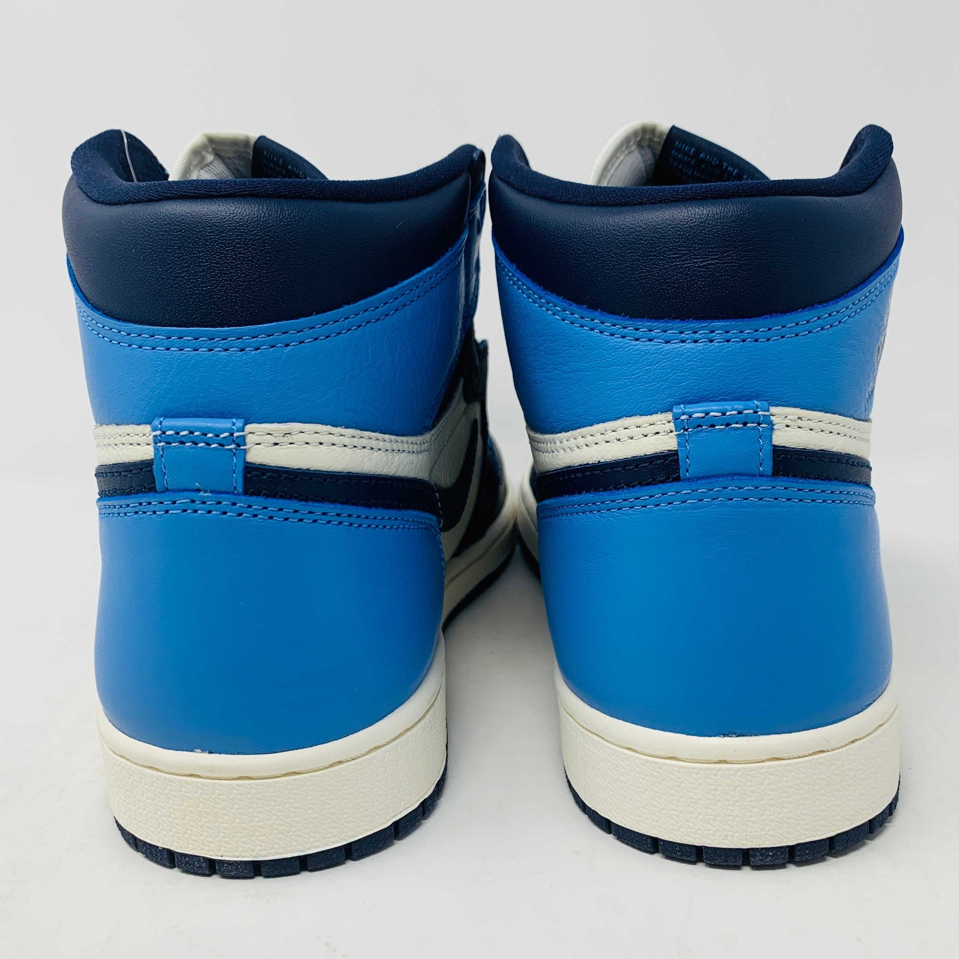 The new Jordan 1 Obsidian high-top sneaker, in blue and white with a black swoosh and blue laces, is displayed on a black and red box featuring 1985 Air Jordan 1 text. A printed Air Jordan wings logo is near the ankle.