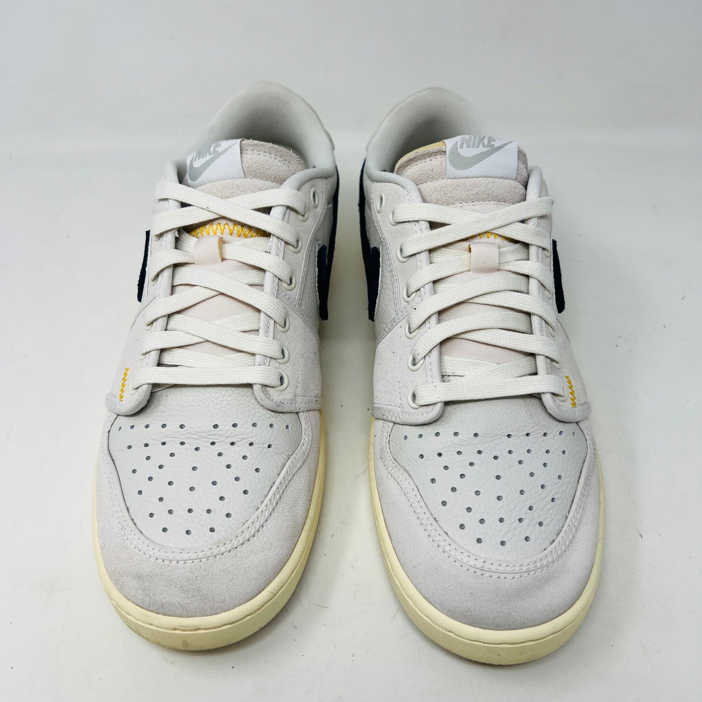 The Jordan 1 Retro AJKO Low SP Union Sail Leather sneakers feature black swooshes, subtle yellow stitching, and white laces. Pictured on a pristine white background, they include extra laces for versatile styling.