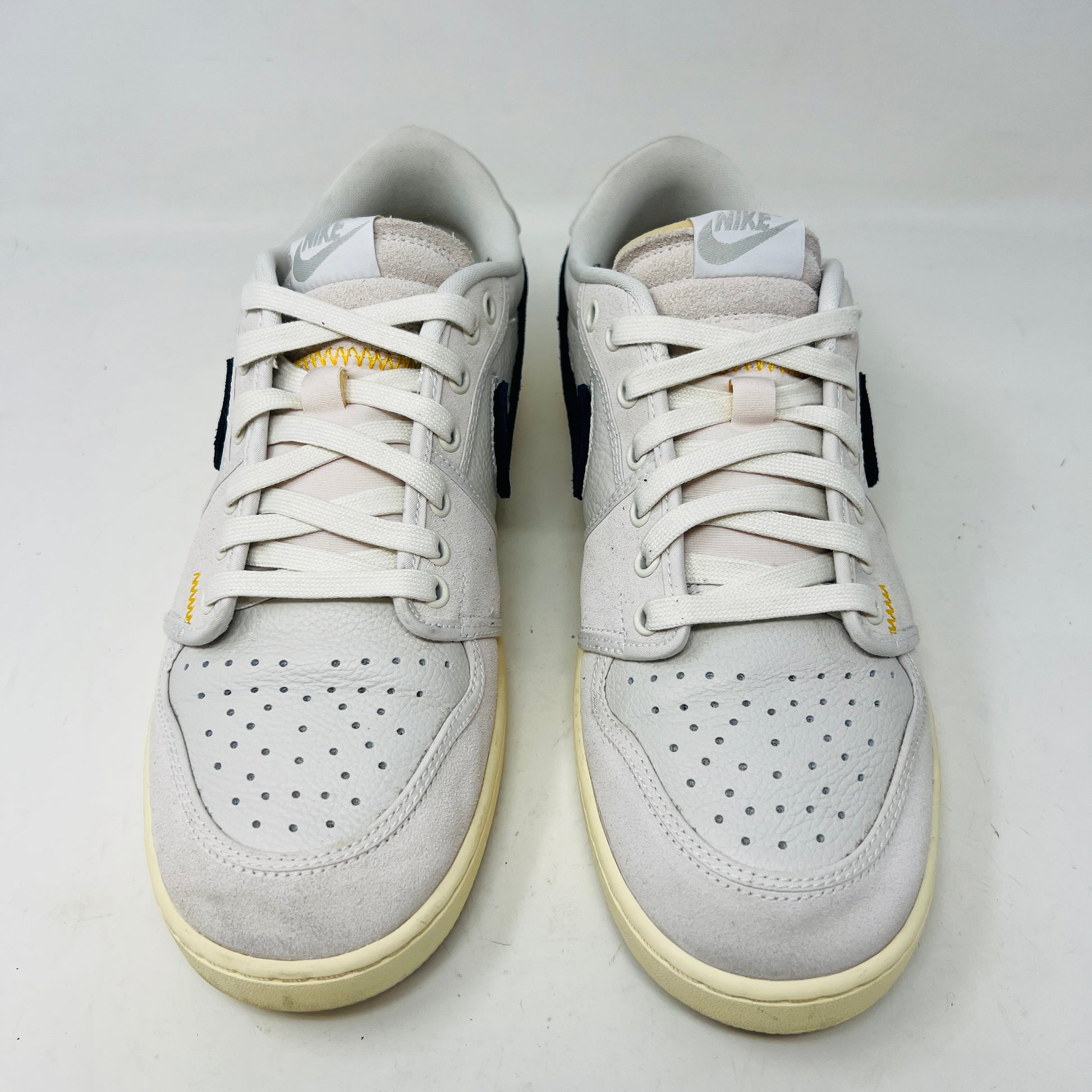 The Jordan 1 Retro AJKO Low SP Union Sail Leather sneakers feature black swooshes, subtle yellow stitching, and white laces. Pictured on a pristine white background, they include extra laces for versatile styling.