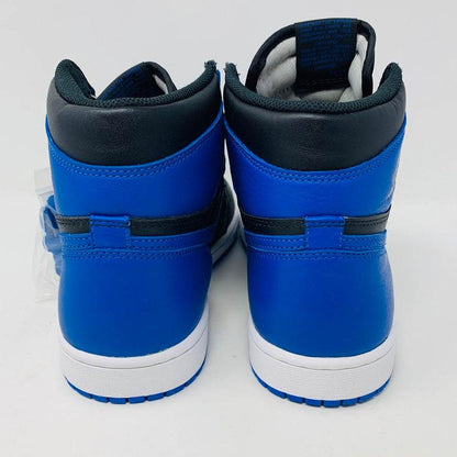 A pair of brand new black and blue Jordan 1 Royal 2017 high-tops are showcased on a black and red Nike box, with a small plastic bag holding authentic blue laces attached to one shoe.