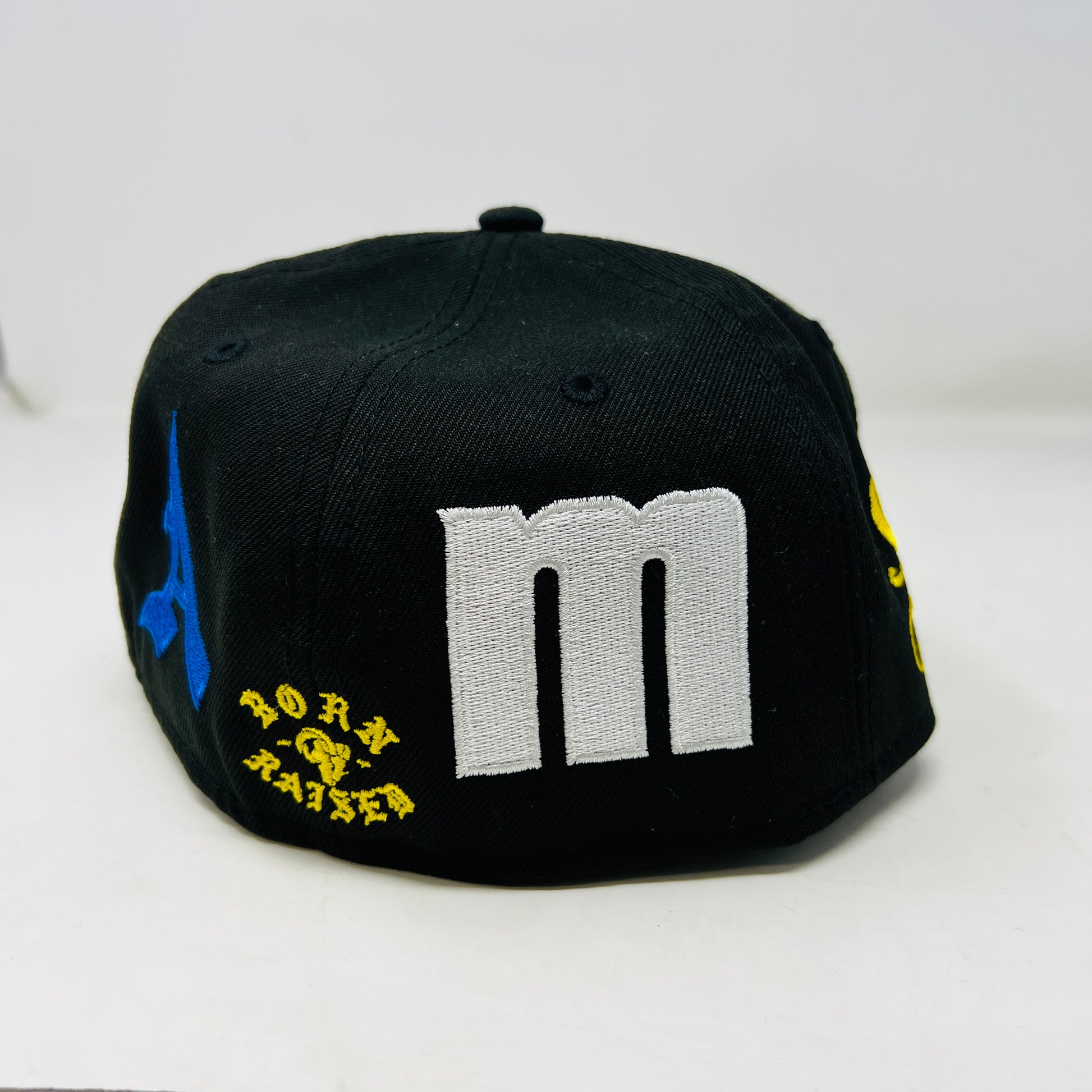 The New Era x Born x Raised x LA RAMS baseball cap by born x raised features LA in yellow and white embroidery on the front. It has a size 7 1/2 sticker on the brim with yellow and blue side patches, all set against a plain white background for a bold statement.