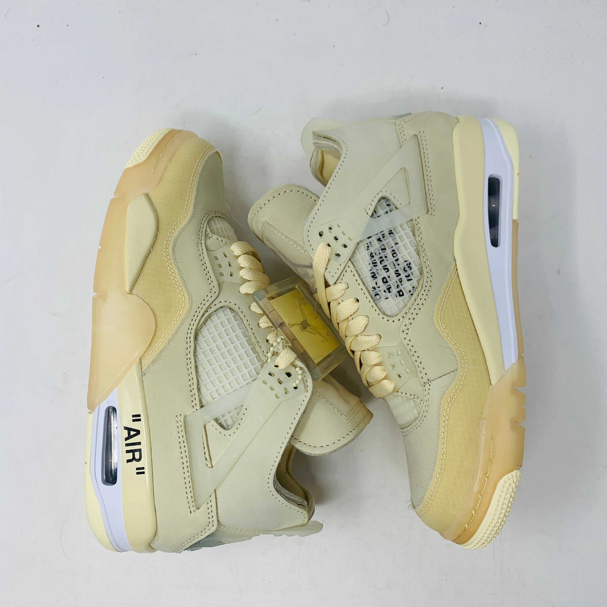 A beige Jordan 4 Off White Sail W sneaker rests on a box with the authentic Air Jordan logo. It comes with three extra sets of laces in mint, cream, and black labeled SHOELACES, a plastic zip tie, and features a small Jumpman Air tag.