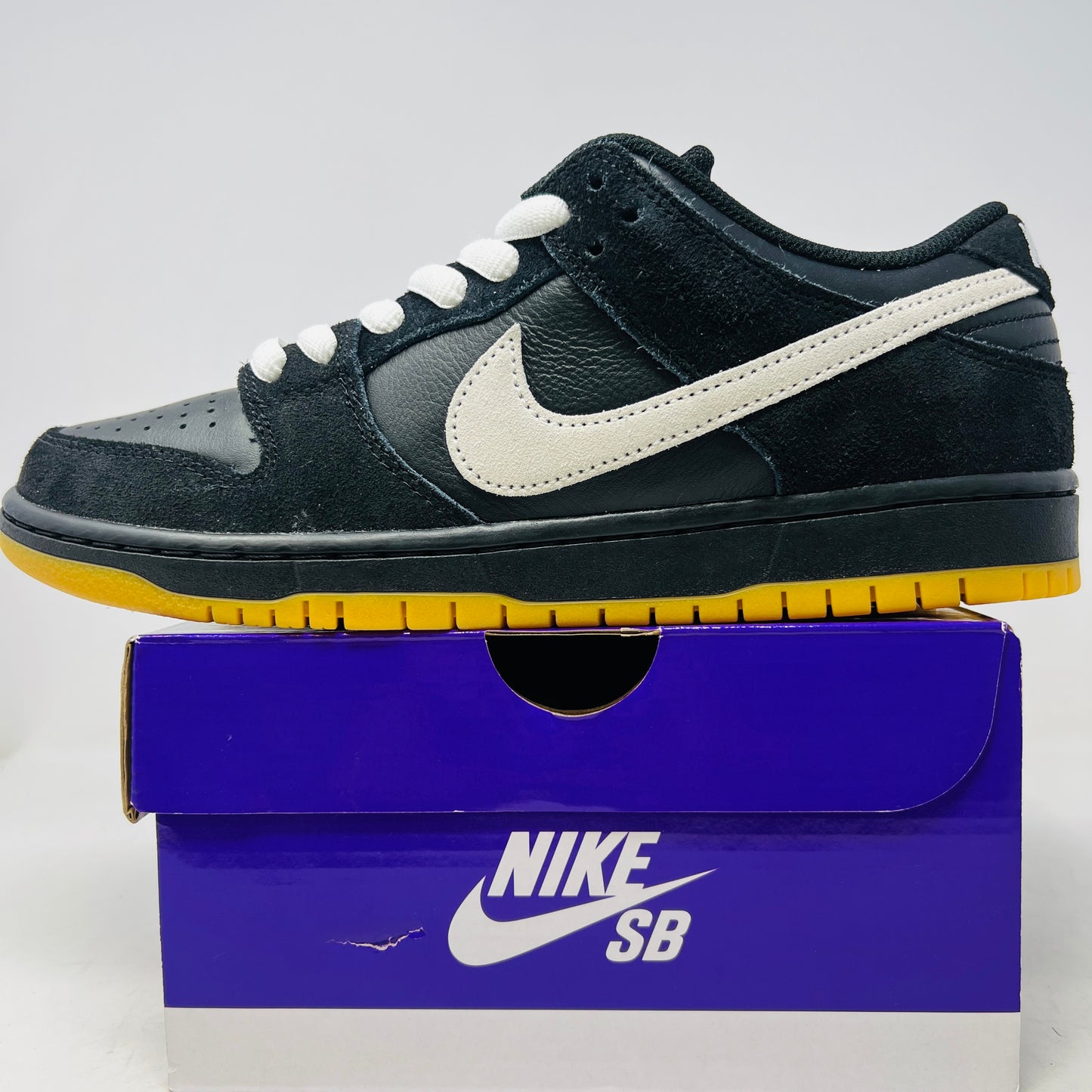 A black Nike Dunk Low Pro SB Black White Gum sneaker with white laces and swoosh sits on a purple Nike SB box. The 2025 shoe has a yellow sole, leather-texture finish, and both are in new condition.