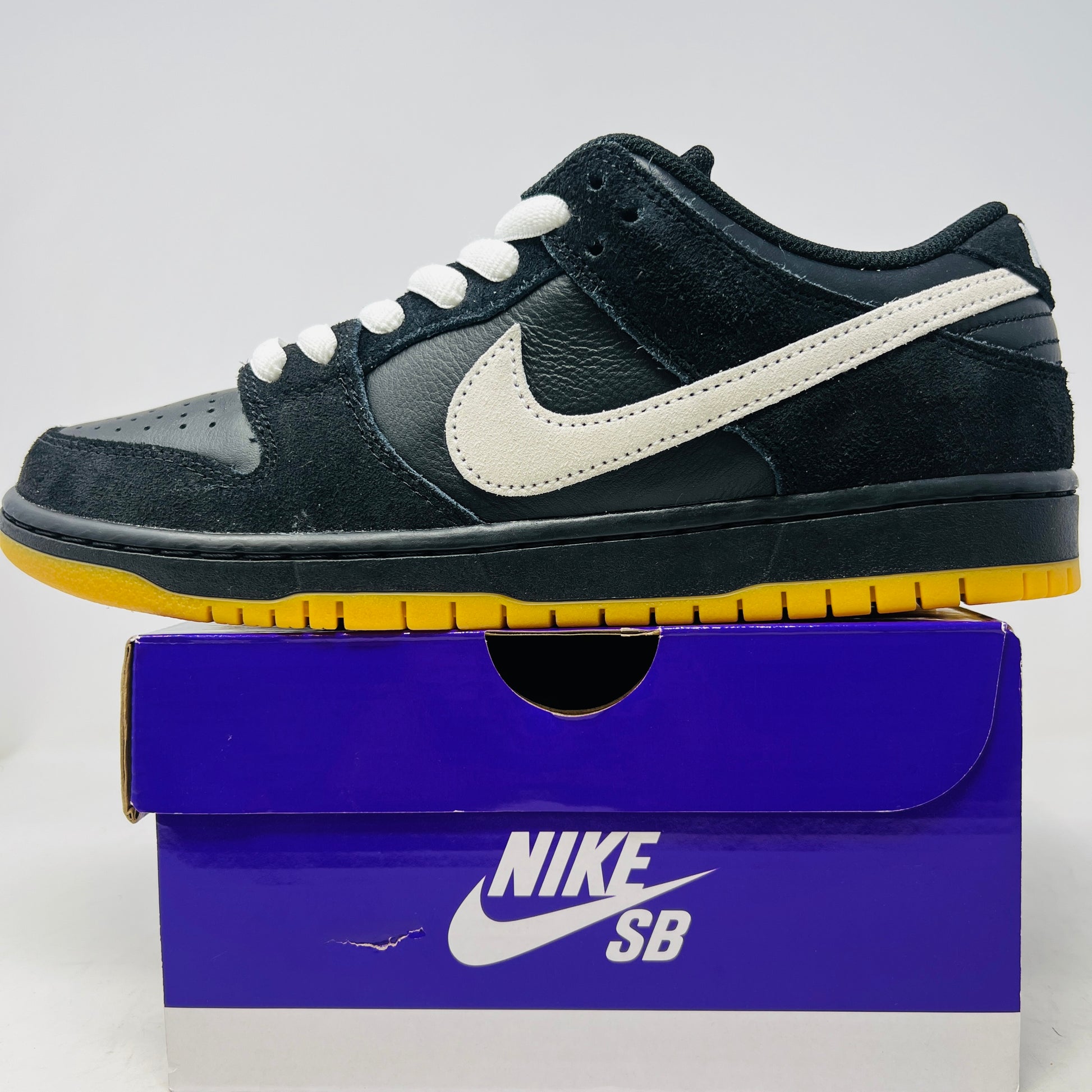 A black Nike Dunk Low Pro SB Black White Gum sneaker with white laces and swoosh sits on a purple Nike SB box. The 2025 shoe has a yellow sole, leather-texture finish, and both are in new condition.