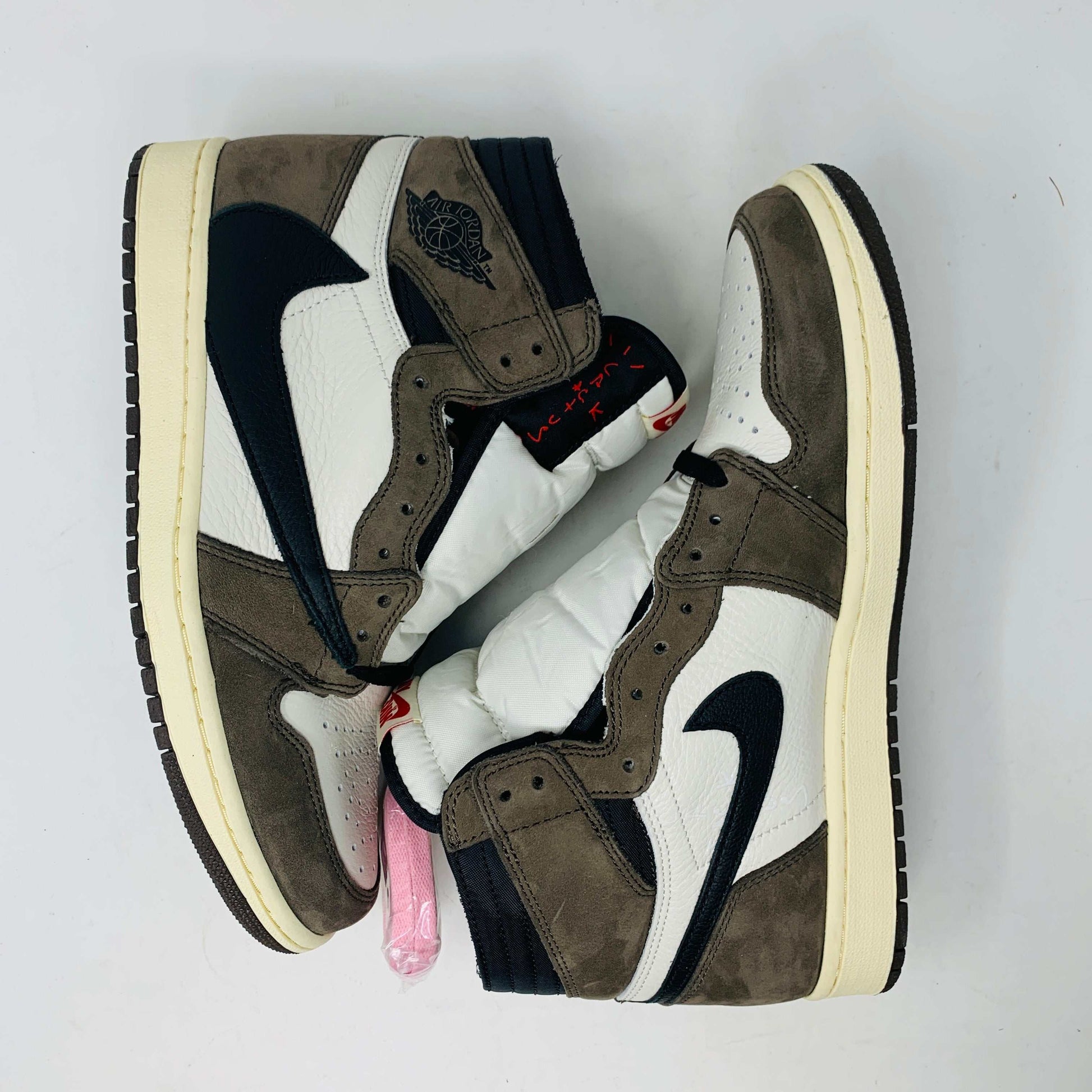 A pair of authentic Jordan 1 High Travis Scott Mocha sneakers in brown, white, and black is displayed on an orange and brown Nike shoebox. These feature a distinctive reversed black Swoosh and come with extra laces for versatile styling.