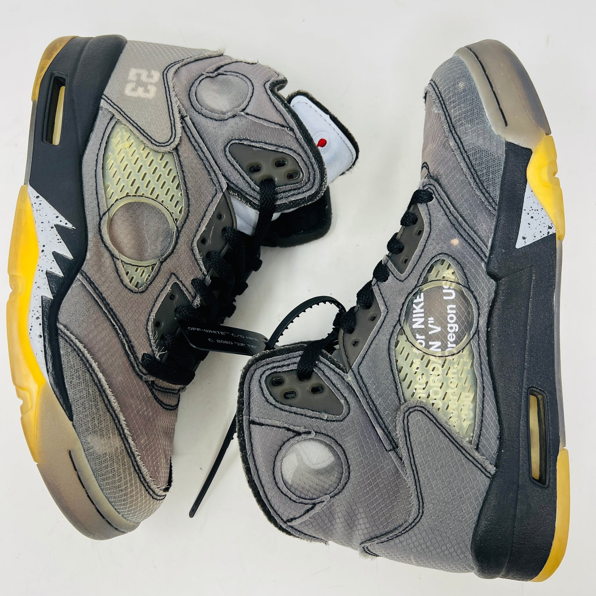 The Jordan 5 Retro Off-White Muslin high-top sneaker, featuring a transparent mesh window and 23 on the heel, is displayed on its box with a white and red shoelace packet in front. A small bleach stain adds unique character to the design.